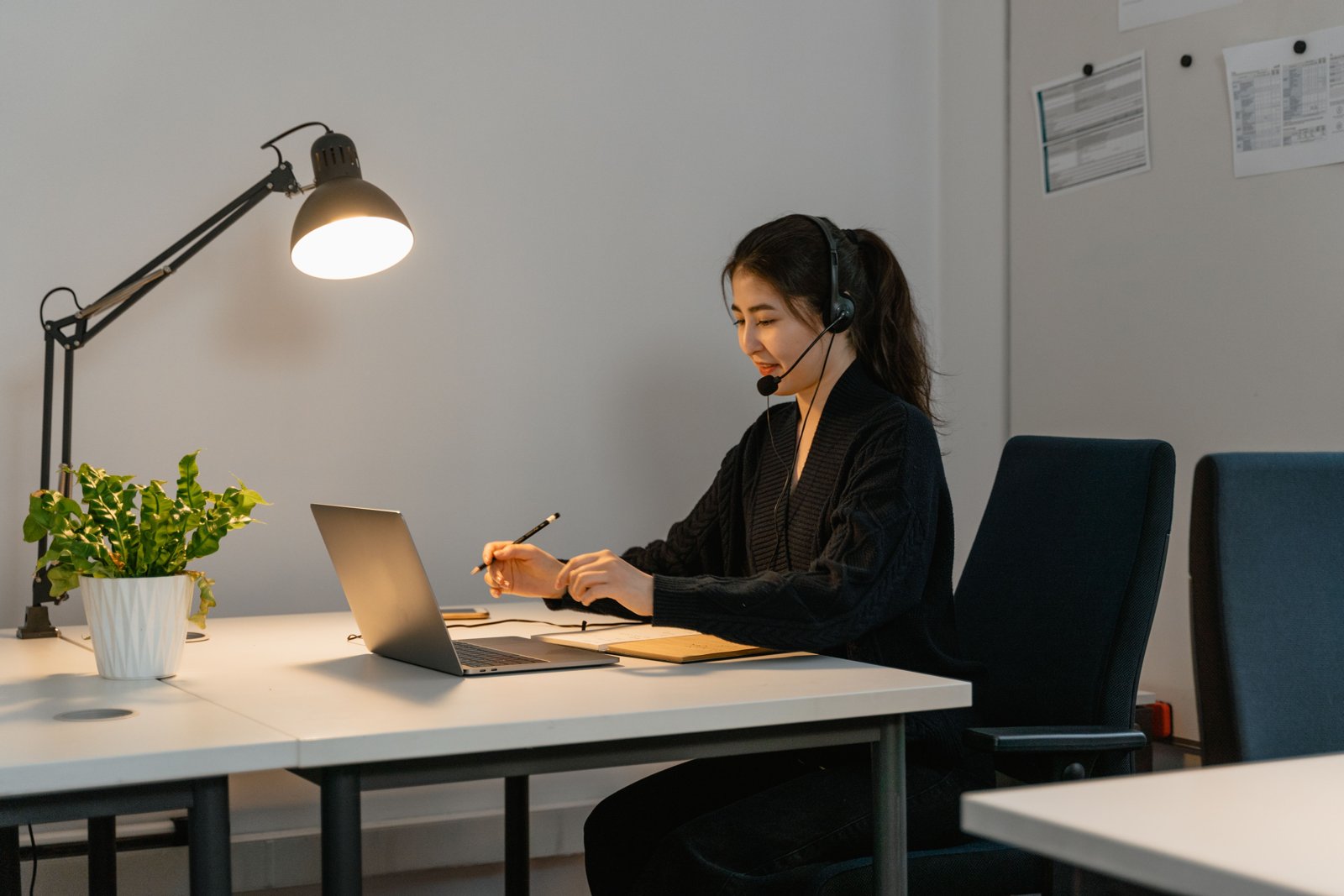 Mulher com headset diante de um notebook, com caderno aberto sobre a mesa.