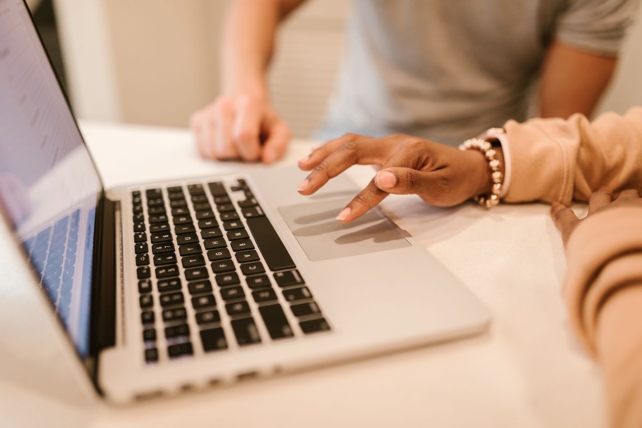 Duas pessoas diante do mesmo notebook, com foco nas mãos de uma delas sobre o touchpad.